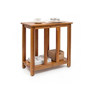May include: A rectangular wooden side table with a warm brown finish, featuring four straight legs and a lower shelf. The top surface holds a white ceramic cup, an open book, and black eyeglasses. A stack of magazines and a woven basket sit below.