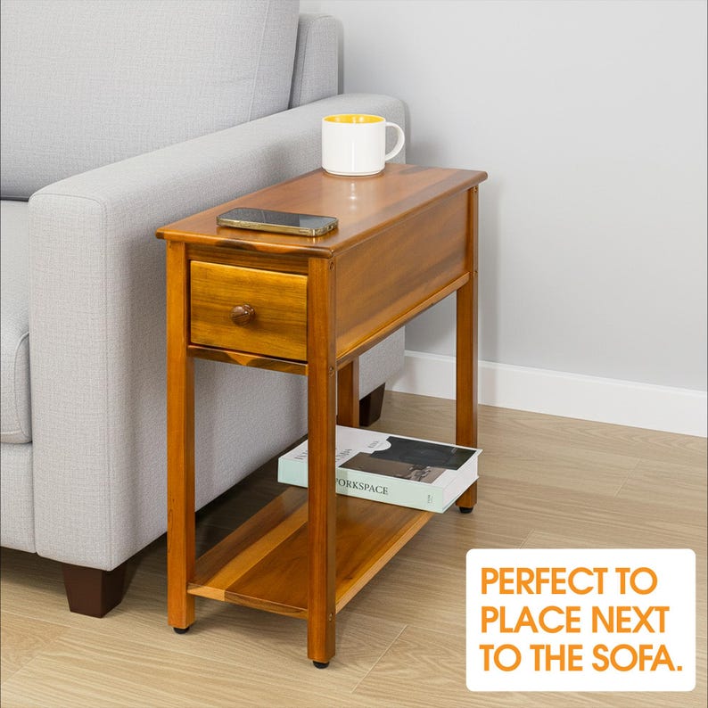May include: A wooden side table with a drawer and a lower shelf. The table is a warm brown color and has a small drawer with a round knob. A white mug with a yellow interior and a smartphone sit on top. The text on the image says "PERFECT TO PLACE NEXT TO THE SOFA."