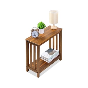 May include: A rectangular brown wooden side table with a slatted side design and a lower shelf. The top surface holds a silver analog alarm clock, a small potted green plant, and a beige lamp. Two books are stacked on the lower shelf.