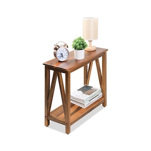 May include: A rectangular wooden console table with an X-frame design on the sides and a lower shelf. On the top surface sits a silver analog alarm clock, a small potted green plant, and a beige lamp. Two books are stacked on the lower shelf.