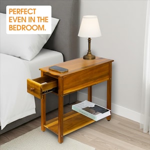 May include: A wooden nightstand with a drawer and a lower shelf. A lamp with a white shade sits on top. The nightstand has a warm, brown finish. The text "PERFECT EVEN IN THE BEDROOM" is in the upper left corner.