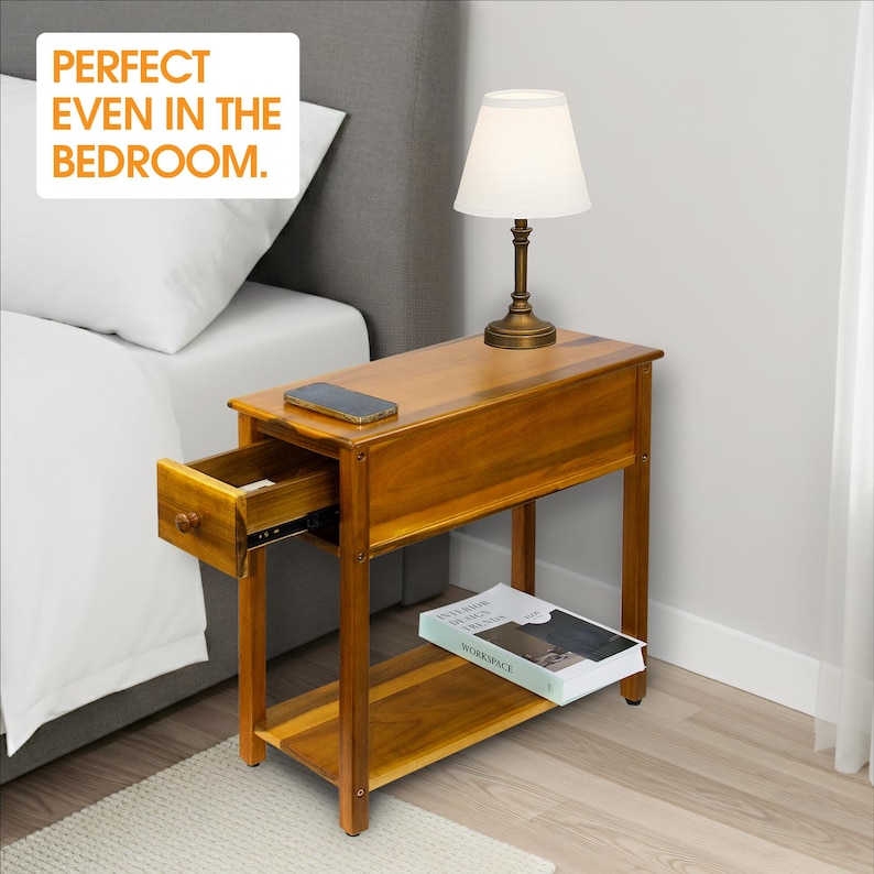 May include: A wooden nightstand with a drawer and a lower shelf. A lamp with a white shade sits on top. The nightstand has a warm, brown finish. The text "PERFECT EVEN IN THE BEDROOM" is in the upper left corner.