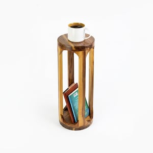 May include: A cylindrical wooden side table with a natural grain finish, featuring an open frame design with vertical supports. A white ceramic mug sits on the top surface, and two books are positioned upright on the bottom shelf.