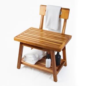 May include: A wooden shower bench with a slatted seat and backrest. A white towel hangs from the back. The bench has a lower shelf holding folded white towels and two bottles. The wood has a warm, natural finish.