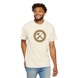 May include: Cream-colored t-shirt featuring a circular graphic with the text "DAD. FIXING THINGS SINCE FOREVER." The graphic includes a hammer and a crossed screwdriver. The person is wearing blue jeans.