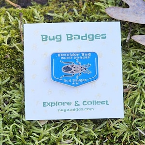 May include: A blue enamel pin featuring a Boxelder Bug, with the scientific name Boisea trivittata, set against a white card. The card also displays the text "Bug Badges" and "Explore & Collect". The pin is on a bed of green moss.