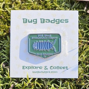 May include: A green and blue enamel pin featuring a pill bug design, labeled "Pill Bug Armadillidium vulgare." The pin is on a white card with the text "Bug Badges" and "Explore & Collect bugbadges.com."