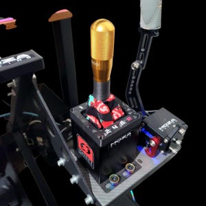 Puede incluir: Primer plano de un conjunto de palanca de cambios de simulador de carreras. La palanca tiene un pomo dorado y una base negra con el logotipo "MOZA Racing". La funda de la palanca tiene un patrón rojo y negro.