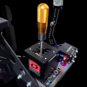 Könnte beinhalten: Ein schwarzer und goldener Rennsimulator-Schalthebel mit dem Text "MOZA Racing" und "KRAFT WORKS". Der Schalthebel hat einen goldenen Griff und einen roten Haken. Das Gerät ist auf einer schwarzen Basis montiert.
