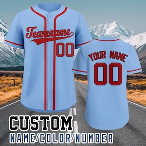 May include: Light blue baseball jersey with red accents and the text "Teamname" and the number "00" on the front. The back of the jersey has the text "YOUR NAME" and the number "00". The bottom of the image has the text "CUSTOM NAME/COLOR/NUMBER".