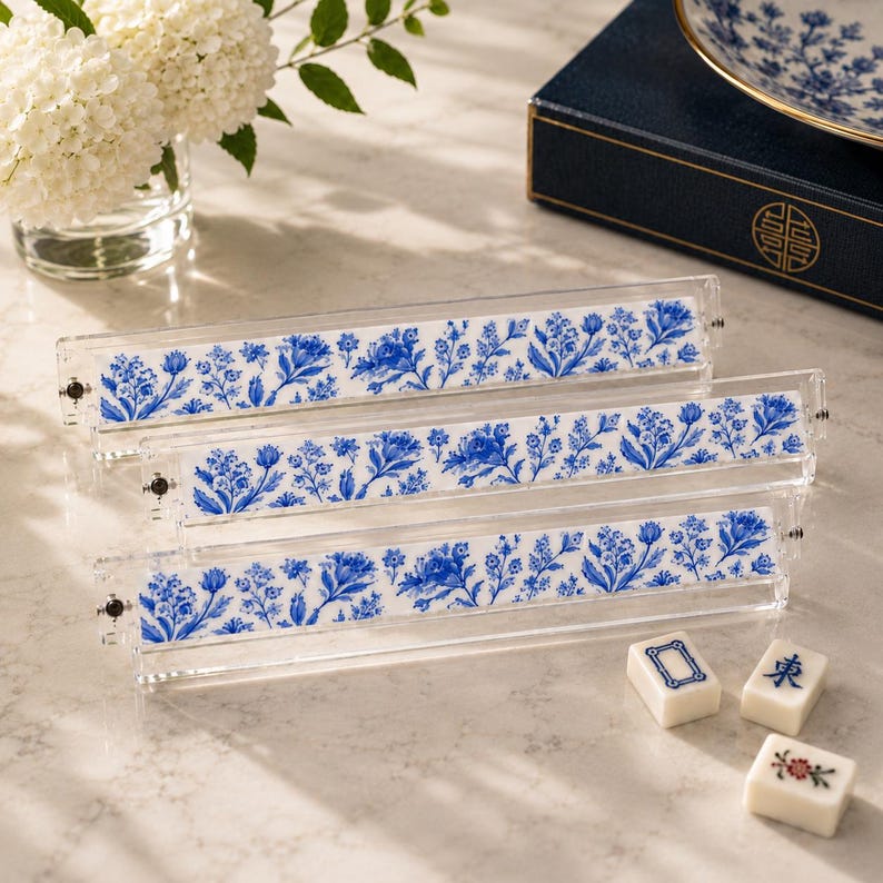 May include: Three clear acrylic mahjong tile racks featuring a decorative insert with a blue and white floral pattern. The racks are arranged in a tiered formation, and three individual mahjong tiles are placed on the surface nearby.