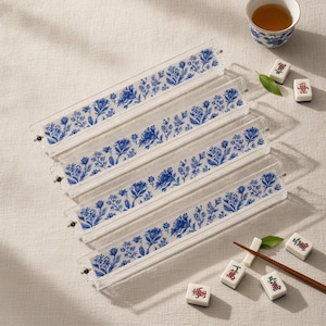 May include: Four clear acrylic mahjong tile racks featuring a blue and white floral pattern. Each rack is rectangular and elongated, designed to hold game tiles. Scattered nearby are several individual mahjong tiles and a wooden stick.