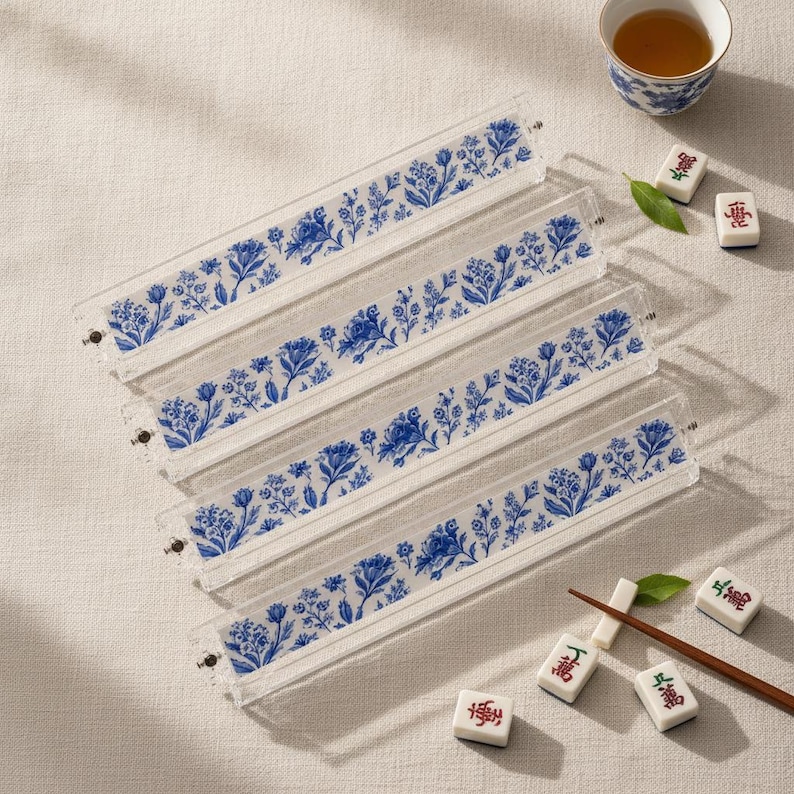 May include: Four clear acrylic mahjong tile racks featuring a blue and white floral pattern. Each rack is rectangular and elongated, designed to hold game tiles. Scattered nearby are several individual mahjong tiles and a wooden stick.