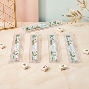 May include: Four clear acrylic mahjong tile racks featuring a tropical palm tree and beach landscape design, accompanied by five individual white mahjong tiles with black and red characters, arranged on a light-colored surface.