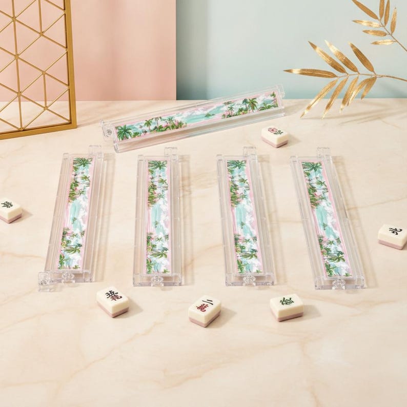 May include: Four clear acrylic mahjong tile racks featuring a tropical palm tree and beach landscape design, accompanied by five individual white mahjong tiles with black and red characters, arranged on a light-colored surface.