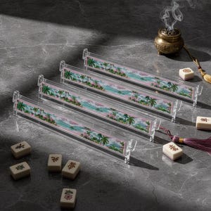 May include: Four clear acrylic mahjong tile racks featuring a colorful tropical landscape print with palm trees and ocean waves. Several ivory-colored mahjong tiles with black and red symbols are scattered nearby, alongside a small incense burner.
