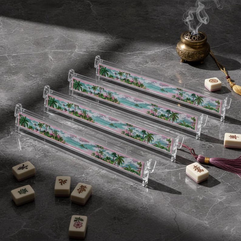 May include: Four clear acrylic mahjong tile racks featuring a colorful tropical landscape print with palm trees and ocean waves. Several ivory-colored mahjong tiles with black and red symbols are scattered nearby, alongside a small incense burner.