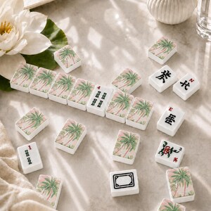May include: A collection of rectangular mahjong tiles featuring a pink and green palm tree pattern on the back. Some tiles display black Chinese characters, red numbers, or floral motifs on their faces. The tiles are white with smooth edges.