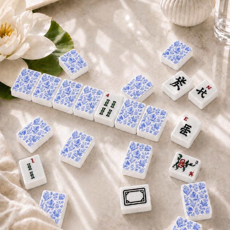 May include: A collection of rectangular mahjong tiles featuring a blue and white floral pattern on the back and various black, red, and green character, number, and symbol designs on the front, scattered across a light-coloured surface.