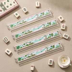 May include: Four clear acrylic mahjong tile racks featuring a printed tropical beach scene with palm trees, turquoise water, and pink sand. Scattered around the racks are individual white mahjong tiles with various green, red, and black symbols.