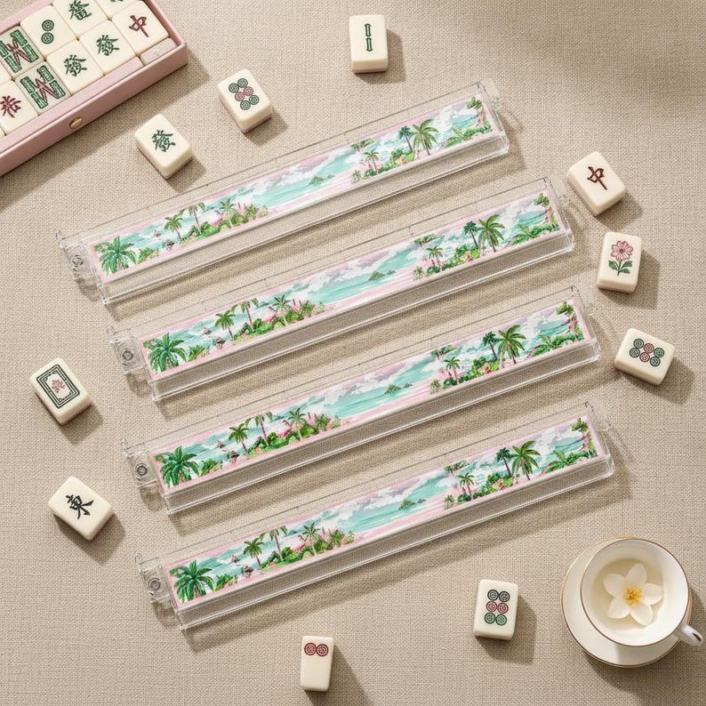May include: Four clear acrylic mahjong tile racks featuring a printed tropical beach scene with palm trees, turquoise water, and pink sand. Scattered around the racks are individual white mahjong tiles with various green, red, and black symbols.