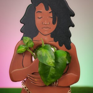 May include: A terracotta-colored wooden planter shaped like a woman with closed eyes, holding a green plant with large leaves. The figure has black hair and a gold chain around the waist. The base is brown, and the logo "Stayplntd" is visible.