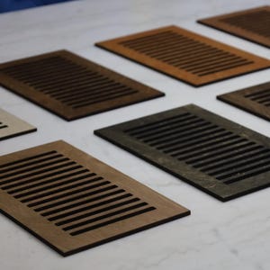 Può includere: Una collezione di coperture per bocchette di ventilazione rettangolari in legno in varie tonalità di marrone, dal beige chiaro all'espresso scuro. Ogni copertura presenta una serie di fessure orizzontali parallele. Le coperture sono disposte su una superficie bianca.