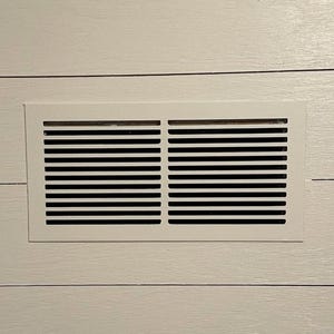 Op de afbeelding: Een witte rechthoekige ventilatieopening met zwarte horizontale lamellen is gecentreerd op een lichtgekleurde, horizontale plankmuur. De muur heeft een houtnerftextuur. Bovenop zit een witte lijst.