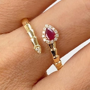 Può includere: Un anello d'oro con una gemma di rubino a forma di pera, circondata da piccoli diamanti. L'anello presenta un design unico con una fascia strutturata che avvolge il dito.