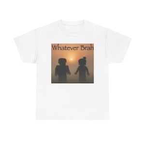 Pode incluir: Uma t-shirt branca de manga curta com uma impressão gráfica central de duas figuras em bloco em silhueta contra um pôr do sol. Acima das figuras, o texto "Whatever Brah" está impresso numa fonte serifada escura e estilizada.