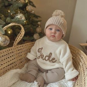 May include: A baby in a cream-coloured knitted sweater embroidered with the name "Jude" in brown, with matching trousers and a beanie hat with a pom-pom. The baby sits in a woven basket, with a Christmas tree visible in the background.