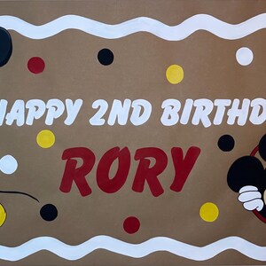 May include: A rectangular brown banner with a white scalloped border featuring Mickey Mouse on the left and a Mickey Mouse head icon on the right. The text reads HAPPY 2ND BIRTHDAY RORY in white and red, accented by scattered red, yellow, black, and white dots.