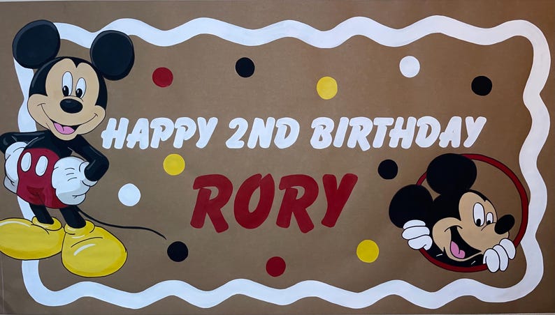 May include: A rectangular brown banner with a white scalloped border featuring Mickey Mouse on the left and a Mickey Mouse head icon on the right. The text reads HAPPY 2ND BIRTHDAY RORY in white and red, accented by scattered red, yellow, black, and white dots.