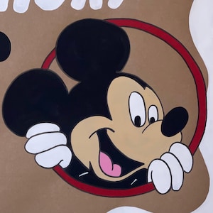 May include: A hand-painted illustration of Mickey Mouse on a tan background. The character features black ears, a tan face, a black nose, and a pink tongue, framed by a thin red circle. White gloved hands are visible at the bottom of the circle.