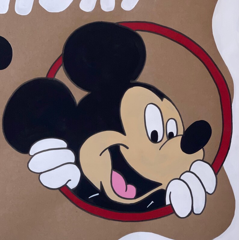 May include: A hand-painted illustration of Mickey Mouse on a tan background. The character features black ears, a tan face, a black nose, and a pink tongue, framed by a thin red circle. White gloved hands are visible at the bottom of the circle.