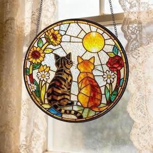 Puede incluir: Un atrapasoles circular de vidrieras que muestra dos gatos sentados uno al lado del otro, uno atigrado y otro naranja, rodeados de girasoles amarillos, margaritas blancas y una rosa roja bajo un sol amarillo brillante, enmarcado en un borde de metal negro.