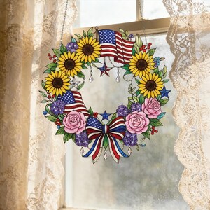 Könnte beinhalten: Ein dekorativer Kranz mit amerikanischen Flaggen, Sonnenblumen, rosa Rosen und lila Blumen. Der Kranz ist mit einer rot-weiß-blauen Schleife und sternförmigen Akzenten verziert. Er ist zum Aufhängen im Fenster gedacht.
