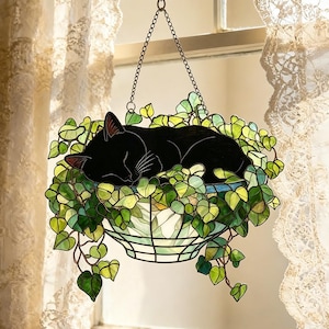 May include: Stained glass hanging planter with a sleeping black cat surrounded by green and yellow ivy. The planter has a geometric design with a blue rim, suspended by a chain. A decorative piece for cat lovers, suitable for a window.