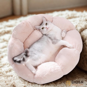May include: A light pink, plush pet bed with a flower-like design. A gray and white cat is lying comfortably inside the bed. The bed has a soft, fluffy texture, and the cat is relaxed. The cat's eyes are open, and it is looking up.