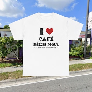 May include: White t-shirt with the text "I ❤️ CAFÉ BÍCH NGA" in black and red. Below the cafe name is the address "5572 Park Blvd, Pinellas Park, FL". The shirt is displayed outdoors with a cafe in the background.