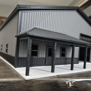 May include: A scale model of a grey and black metal building featuring a front porch with black support pillars, a central door, two windows, and a large garage door opening on the side. The structure sits on a white rectangular base.