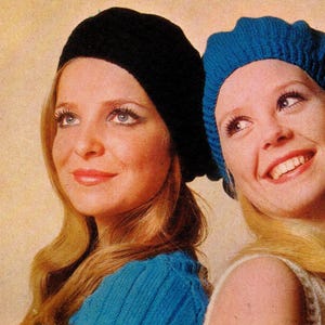 May include: Two women wearing knitted beret hats. One hat is black, the other is blue. Both women have long blonde hair and are smiling. The image has a vintage, retro aesthetic.