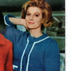 Op de afbeelding: Een vrouw draagt een blauw vest met witte details en knopen. Het vest heeft twee voorzakken en wordt gecombineerd met een witte rok. De afbeelding komt uit een vintage tijdschrift.