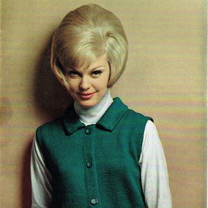 Può includere: Una donna indossa un abito in maglia verde con un gilet abbottonato con colletto sopra un dolcevita bianco e una camicia a maniche lunghe. La donna ha i capelli biondi e sorride.