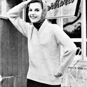 以下が含まれることがあります： 明るい色のVネックセーター、柄のスカーフ、濃い色のズボンを着た人物の白黒写真。その人は笑顔で木の構造物に寄りかかっています。画像はレトロな美学を持っています。