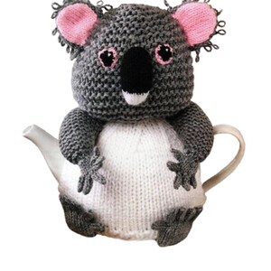 May include: A knitted tea cosy in the shape of a koala bear. The cosy is grey and white, with pink ears and a black nose. The koala design is wrapped around a white teapot, with the arms and feet of the koala visible.
