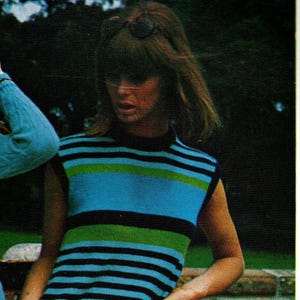 May include: A sleeveless, knee-length dress with horizontal stripes in blue, green, and black. The dress has a high neckline and is worn by a person leaning against a wooden railing. The overall style is retro.