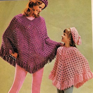 Può includere: Una donna e una bambina indossano poncho e cappelli all'uncinetto abbinati. Il poncho della donna è viola scuro con frange, e quello della bambina è rosa chiaro. Entrambi gli outfit hanno uno scollo a V.
