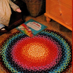 May include: A handmade, circular rug with a vibrant, rainbow-colored design. The rug features concentric rings of color, transitioning from a warm orange center to outer rings of red, blue, and black. A Dr. Seuss book rests on the rug.