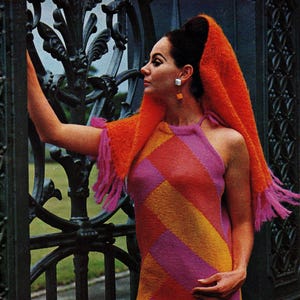 May include: A vintage halter dress with a geometric pattern in orange, pink, and yellow. The dress is worn with an orange shawl with pink fringe. The woman is near a black wrought iron gate. The dress is knee-length.